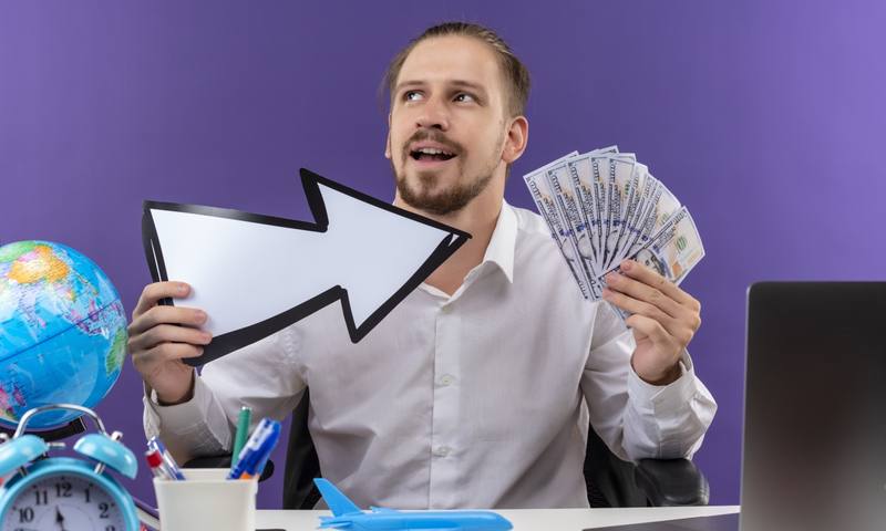 handsome-businessman-white-shirt-holding-white-arrow-showing-cash-looking-aside-with-smile-face-sitting-table-offise-purple-background