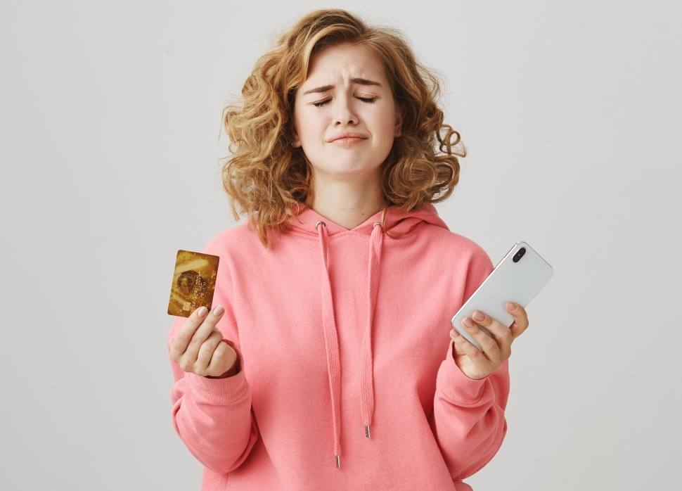 upset-curly-haired-girl-frowning-lacking-money-bank-account-hold-mobile-phone
